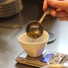 Une main verse des herbes séchées dans une tasse blanche posée sur une balance. Le dosage précis est essentiel pour 1 litre d'infusion.