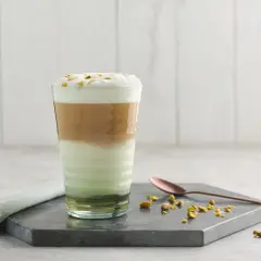 Un latte pistache aux couches distinctes, couronné de mousse et de pistaches concassées. Une cuillère en cuivre repose à côté.