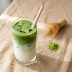 Un verre de pâte minute claire au matcha, glacé et rafraîchissant, avec une paille rose. Un fouet à matcha et une pincée de poudre verte à côté.