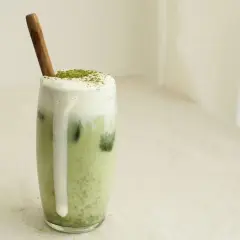 Un verre de matcha lait d'amande crémeux, garni de mousse et d'une pincée de poudre verte, avec une cuillère en bois.