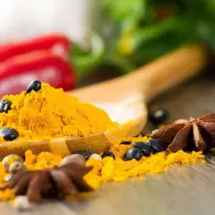 Une cuillère en bois déborde de poudre de curcuma, parsemée de grains de poivre noir et d'anis étoilé. Un **tabel karouia** pour une cuisine pleine de saveurs.