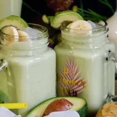 Deux verres de jus d'avocat crémeux, garnis de rondelles de banane et d'avocat, avec des fruits frais en arrière-plan.