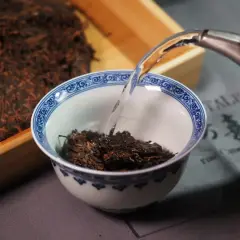 Préparation d'un thé de Chine. De l'eau chaude est versée dans un bol rempli de feuilles de thé sombres, à côté d'un bloc de thé compressé.