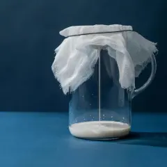 Un pichet en verre contient du kéfir, recouvert d'une étamine blanche retenue par un élastique. La boisson fermentée coule doucement.