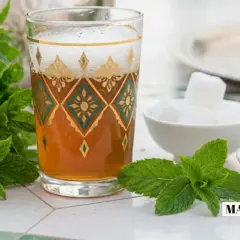 Un verre de thé à la menthe fraîche, orné de motifs dorés et verts, est prêt à être dégusté avec des morceaux de sucre et des feuilles de thé.
