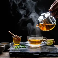 Une main verse un thé rose fumant dans une tasse. Du miel, de la menthe et de la citron vert accompagnent cette boisson réconfortante.