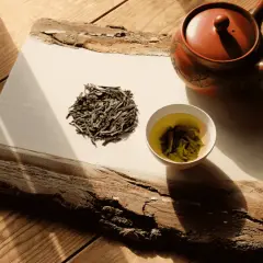 Théière en terre cuite, feuilles de bancha sèches et une tasse de thé vert infusé, sur un plateau en bois brut éclairé par le soleil.