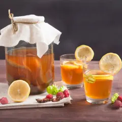 Un grand bocal de boisson kombucha, deux verres garnis de citron et de menthe, avec des framboises et une tranche de citron sur une serviette.