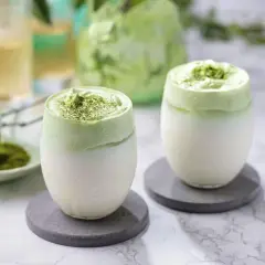 Deux verres de dalgona matcha, une boisson crémeuse et verte, sur des sous-verres gris.