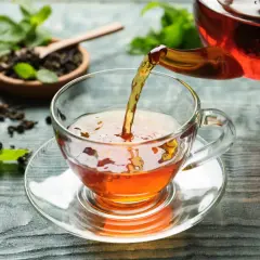 Un thé ambré est versé dans une tasse transparente. Le thé est-il bon pour la santé ? Des feuilles de menthe et du thé en vrac sont disposés sur la table.