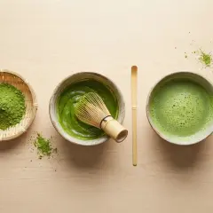 Préparation du matcha : poudre verte dans un panier, pâte dans un bol avec un fouet, et boisson mousseuse dans un autre bol. Le tout sur fond de bois clair.