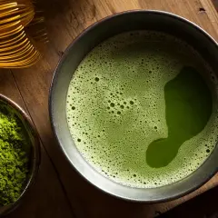Une composition de matcha : poudre verte vibrante dans un bol, une tasse de thé mousseux et un fouet en bambou sur fond de bois.