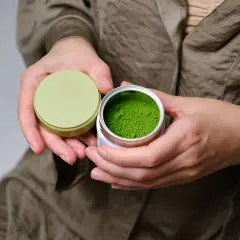 Mains tenant une boîte ouverte de poudre de matcha verte vibrante, évoquant les matcha bienfaits peau.