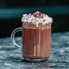 Un réconfortant chocolat chaud chantilly, garni de cacao, dans un mug en verre sur un marbre sombre.