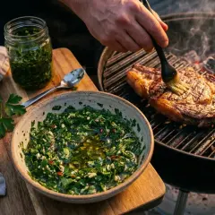 Un steak grillé est badigeonné avec une sauce chimichurri fraîche. Une autre portion de sauce est dans un bol à côté.