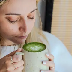Femme aux cheveux blonds savourant un matcha latte crémeux, le regard clos. La boisson verte présente un joli dessin de mousse.