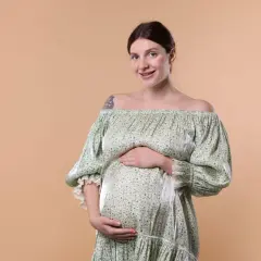 Une femme enceinte, au teint clair et aux cheveux bruns tirés en arrière, sourit à la caméra. Elle porte une robe verte à fleurs, dénudant ses épaules, et pose ses mains sur son ventre arrondi.
