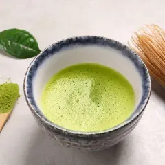 Bol rempli de matcha mousseux, une cuillère en bois avec de la poudre verte et un fouet en bambou. Une feuille de thé verte repose à côté, évoquant le pouvoir antioxydant du matcha.