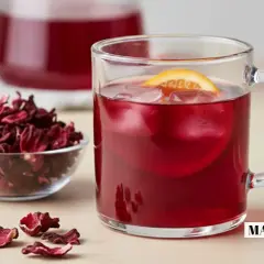 Verre de boisson rouge vif avec glaçons et tranche d'orange, à côté d'un bol de fleurs d'hibiscus séchées. L'eau d'hibiscus, rafraîchissante et colorée.