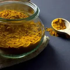 Un pot en verre rempli de poudre de curcuma, avec une cuillère en bois à côté. Ces épices, comme le cumin, apportent couleur et saveur.