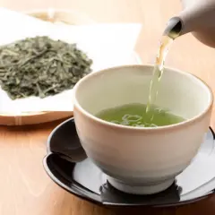 Comment faire du thé vert : une théière verse un liquide vert vif dans une tasse, à côté d'une pile de feuilles de thé séchées.