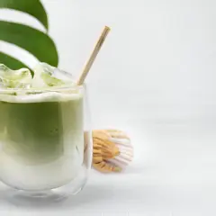 Un verre de thé matcha glacé, parfait pour savoir si on peut boire le thé matcha froid. Une feuille de monstera et un fouet à matcha complètent la scène.