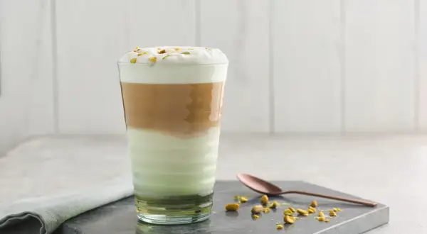 Un latte pistache aux couches distinctes, couronné de mousse et de pistaches concassées. Une cuillère en cuivre repose à côté.