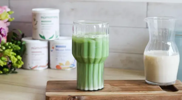 Un verre de matcha chaud, mousseux et crémeux, prêt à être dégusté. À côté, une carafe de lait végétal.
