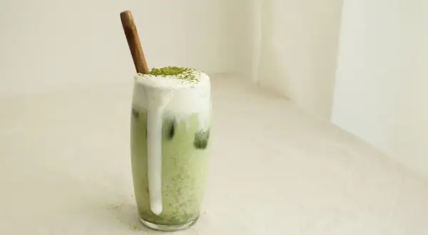 Un verre de matcha lait d'amande crémeux, garni de mousse et d'une pincée de poudre verte, avec une cuillère en bois.
