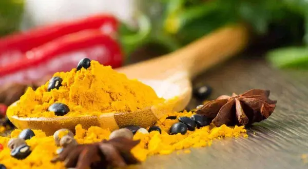 Une cuillère en bois déborde de poudre de curcuma, parsemée de grains de poivre noir et d'anis étoilé. Un **tabel karouia** pour une cuisine pleine de saveurs.