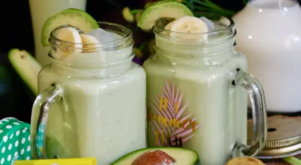Deux verres de jus d'avocat crémeux, garnis de rondelles de banane et d'avocat, avec des fruits frais en arrière-plan.