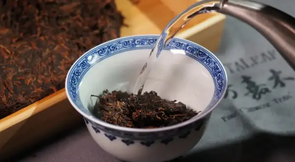 Préparation d'un thé de Chine. De l'eau chaude est versée dans un bol rempli de feuilles de thé sombres, à côté d'un bloc de thé compressé.
