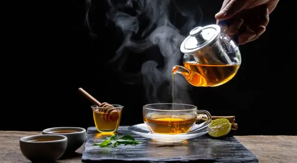 Une main verse un thé rose fumant dans une tasse. Du miel, de la menthe et de la citron vert accompagnent cette boisson réconfortante.