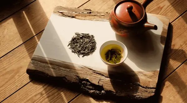 Théière en terre cuite, feuilles de bancha sèches et une tasse de thé vert infusé, sur un plateau en bois brut éclairé par le soleil.