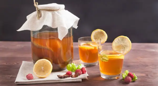 Un grand bocal de boisson kombucha, deux verres garnis de citron et de menthe, avec des framboises et une tranche de citron sur une serviette.
