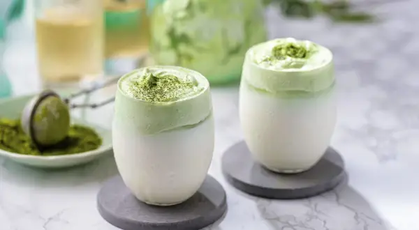 Deux verres de dalgona matcha, une boisson crémeuse et verte, sur des sous-verres gris.