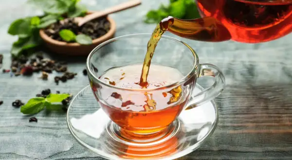 Un thé ambré est versé dans une tasse transparente. Le thé est-il bon pour la santé ? Des feuilles de menthe et du thé en vrac sont disposés sur la table.