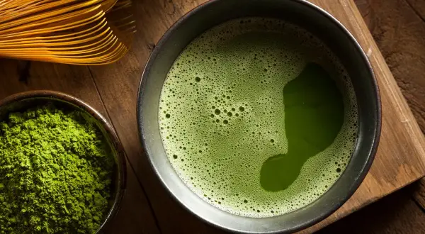 Une composition de matcha : poudre verte vibrante dans un bol, une tasse de thé mousseux et un fouet en bambou sur fond de bois.