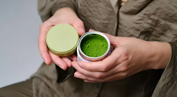 Mains tenant une boîte ouverte de poudre de matcha verte vibrante, évoquant les matcha bienfaits peau.