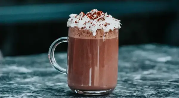 Un réconfortant chocolat chaud chantilly, garni de cacao, dans un mug en verre sur un marbre sombre.