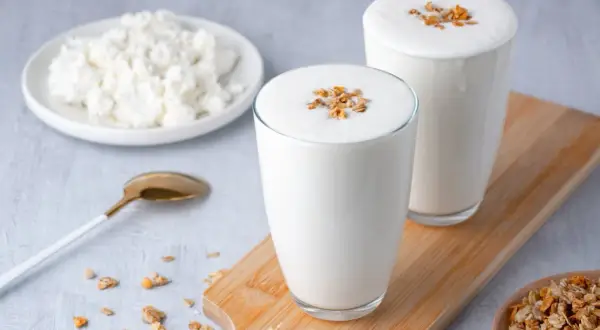 Deux verres de kefir de lait crémeux, garnis de granola, à côté d'une assiette de fromage blanc et d'une cuillère dorée.