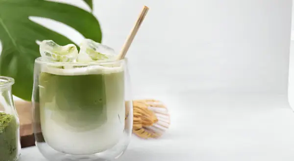 Un verre de matcha à jeun, glacé et crémeux, avec une paille en bois. À côté, un fouet à matcha et une feuille de Monstera.