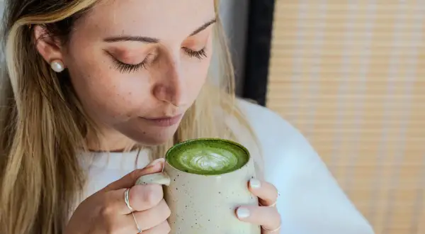 Femme aux cheveux blonds savourant un matcha latte crémeux, le regard clos. La boisson verte présente un joli dessin de mousse.