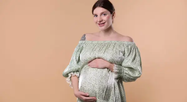 Une femme enceinte, au teint clair et aux cheveux bruns tirés en arrière, sourit à la caméra. Elle porte une robe verte à fleurs, dénudant ses épaules, et pose ses mains sur son ventre arrondi.