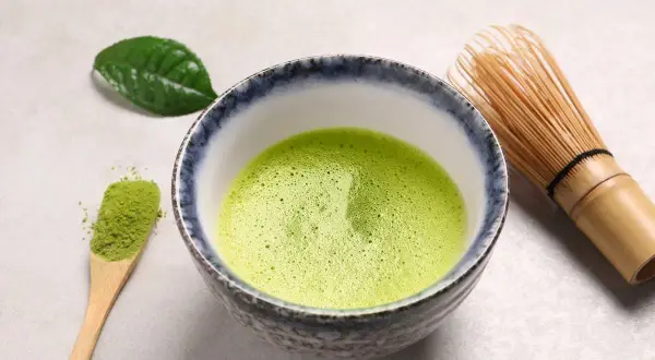 Bol rempli de matcha mousseux, une cuillère en bois avec de la poudre verte et un fouet en bambou. Une feuille de thé verte repose à côté, évoquant le pouvoir antioxydant du matcha.