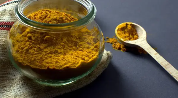 Un pot en verre rempli de poudre de curcuma, avec une cuillère en bois à côté. Ces épices, comme le cumin, apportent couleur et saveur.