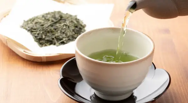Comment faire du thé vert : une théière verse un liquide vert vif dans une tasse, à côté d'une pile de feuilles de thé séchées.