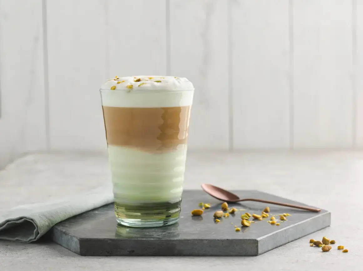 Un latte pistache aux couches distinctes, couronné de mousse et de pistaches concassées. Une cuillère en cuivre repose à côté.