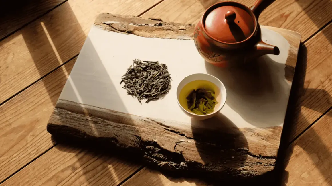 Théière en terre cuite, feuilles de bancha sèches et une tasse de thé vert infusé, sur un plateau en bois brut éclairé par le soleil.