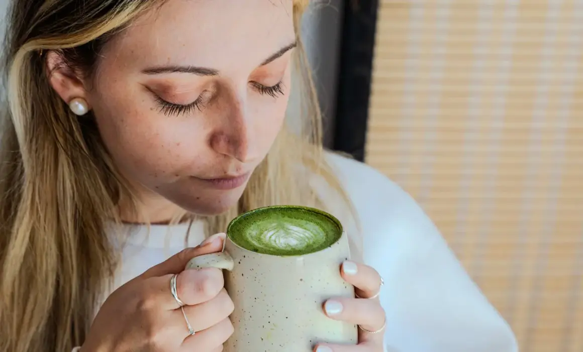 Femme aux cheveux blonds savourant un matcha latte crémeux, le regard clos. La boisson verte présente un joli dessin de mousse.